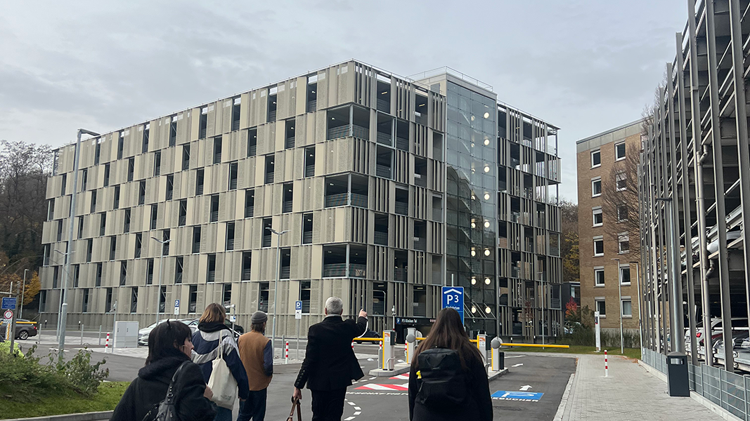 Prof. Dr. Seidl mit einer Gruppe Studierender am unteren Bildrand in der Rückansicht; er zeigt auf das Parkhaus der Kliniken Tal, einen achtstöckigen Bau Stahlbau mit einem eingesetzten gläsernen Treppenhaus