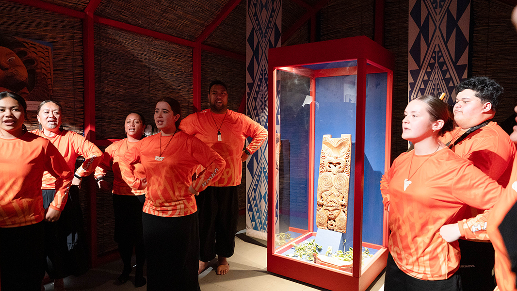 Die Schüler*innen als Vertreter der Te Aitanga-a-Hauiti aus Uawa führen um die beleuchtete Vitrine mit Hinematioros Pou herum einen Tanz auf; alle haben die Hände in die Seiten gestemmt