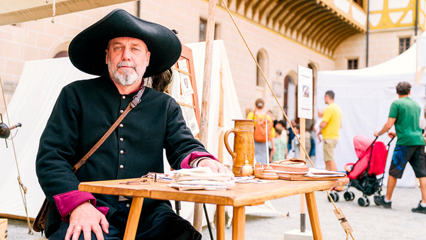 Mann mit schwarzem Uniformrock und schwarzem, ausladendem Schlapphut sitzt an einem niedrigen Holztisch in einem Zelt