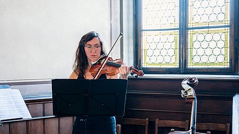Eine Musikerin spielt, durch ein Notenpult zum Teil verdeckt, die Violine vor einem Kirchenfenster