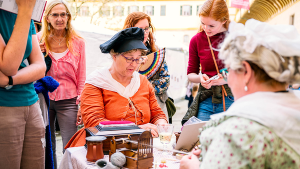 Dame des 18. Jahrhundert sin orangenem Kleid mit Spitzenkragen verrichtet eine Handarbeit, umgeben von beobachtenden Besuchenden