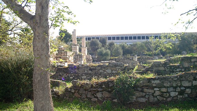 Foto von Ruinen auf der klassischen Agora von Athen, mit einem Baum links im Vordergrund, bewachsenen Mauerresten im Mittelgrund, links mit aufragenden Säulen, und im Hintergrund unscharf die rekontruierte Stoa des Attalos