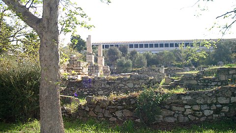 Foto von Ruinen auf der klassischen Agora von Athen, mit einem Baum links im Vordergrund, bewachsenen Mauerresten im Mittelgrund, links mit aufragenden Säulen, und im Hintergrund unscharf die rekontruierte Stoa des Attalos