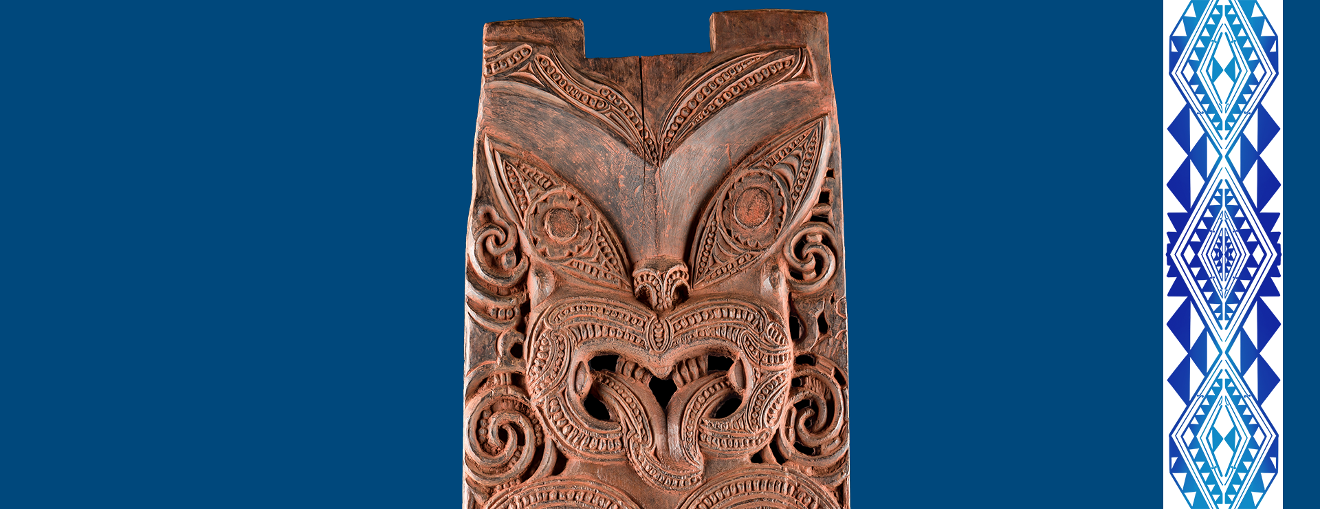 Geschnitztes Holzpaneel (pou) mit der Darstellung einer Ahnin der Te Aitanga-a-Hauiti vor dunkelblauem Hintergrund; rechts ein schmaler Streifen mit einem vor allem aus dreieckigen Formen zusammengesetzten, abstrakten, blauen Design vor weißem Hintergrund