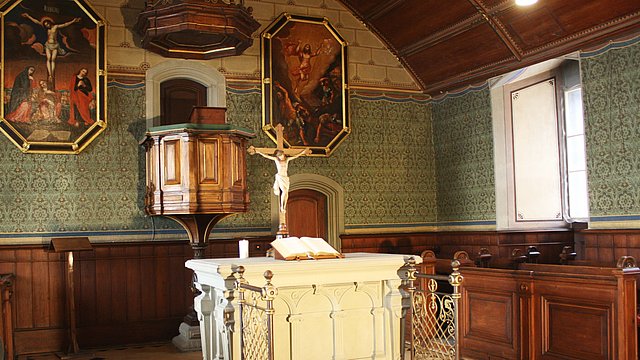 Foto: MUT Blick in den Altarbereich der Schlosskirche mit weißem Altar mit aufgeschlagener Bibel, gekreuzigter Christusfigur und hinaufführender Treppe, im Hintergrund Kanzel, flankiert von Malereien des gekreuzigten (links) und in den Himmel fahrenen Christus (rechts)