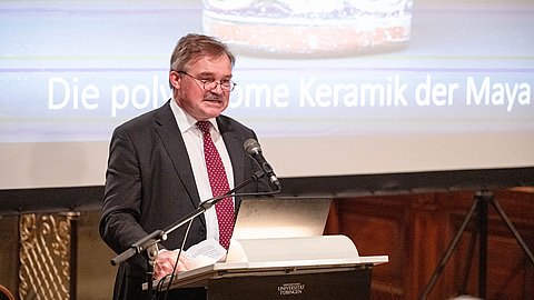 © MUT | Valentin Marquardt Professor Dr. Dr. h. c. Nikolai Grube am Rednerpult am Altarbereich der Schlosskirche Hohentübingen, hinter ihm die Projektion auf der Leinwand mit einem Keramikgefäß und dem Schriftzug "Die polychrome Keramik der Maya"