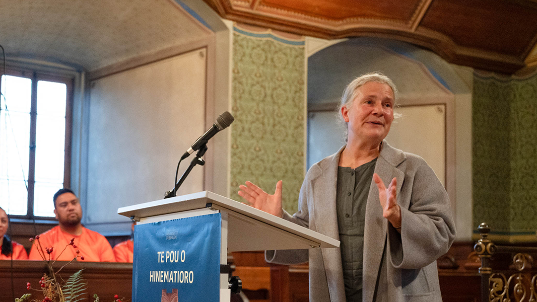 Am Rednerpult mit dem blauen Ausstellungsplakat von "Te Pou o Hinematioro" steht Universitätsrektorin Professorin Dr. Dr. h.c. (Doshisha) Karla Pollmann und wendet sich mit im Sprechen erhobenen Händen nach rechts
