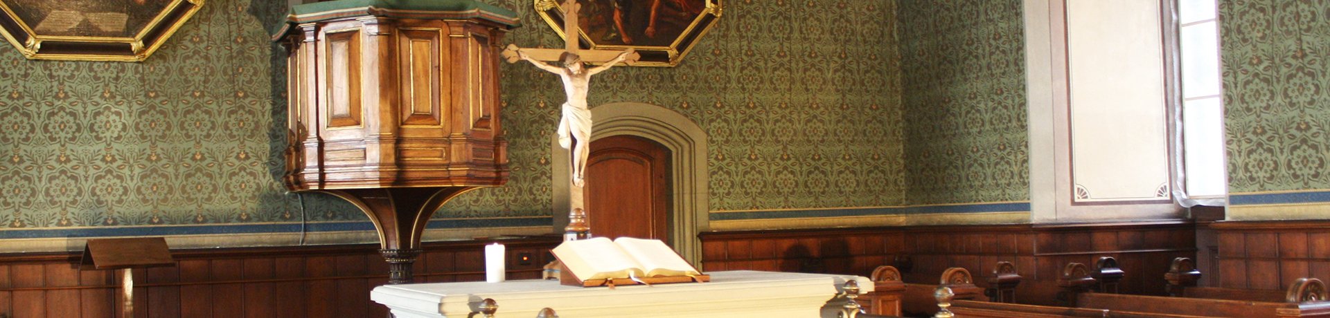 Blick in den Altarbereich der Schlosskirche mit weißem Altar mit aufgeschlagener Bibel, gekreuzigter Christusfigur und hinaufführender Treppe, im Hintergrund Kanzel, flankiert von Malereien des gekreuzigten (links) und in den Himmel fahrenen Christus (rechts)