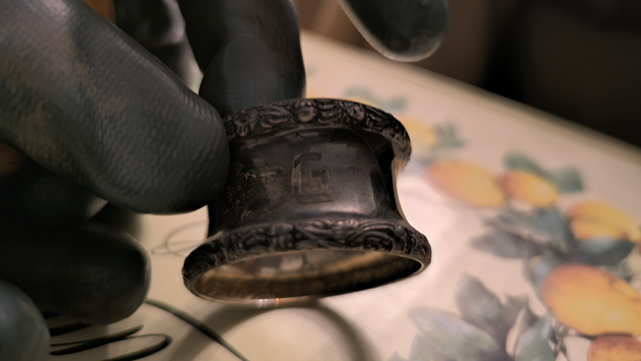 Schwarz behandschuhte Hand hält einen dunkel angelaufenen Metallring mit ornamentierten Rändern