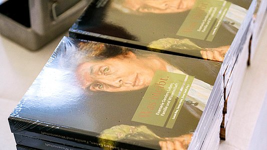 Blick auf zwei Stapel mit Ausstellungskatalogen zu "Was bleibt. Evamarie Sanders Leben, Familie und Wissenschaft" mit Professorinnenportrait von Professorin Sander vor dunklem Hintergrund