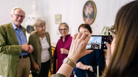 Vor dem Hintergrund des nachgebildeten Biedermeierzimmers wird ein Gruppenfoto von vier Personen gemacht, die auf dem Bildschirm des fotografierenden Handys in der rechten Bildhälfte scharf zu erkennen sind
