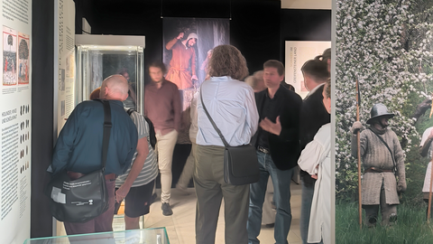 Foto: Michael Kienzle Besuchende stehen in der Ausstellung vor hohen Glasvitrinen mit mehreren Ebenen und blicken auf Wandtafeln; vorne rechts ist der Rand einer freistehenden Fototafel mit einem kostümierten Bewaffneten zu sehen