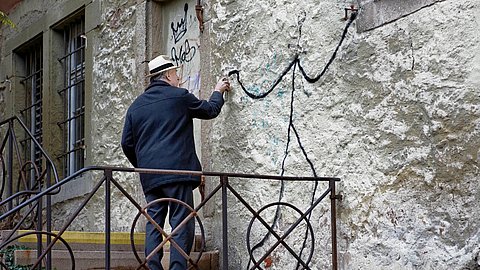 Foto: Charlotte Hermann und Uli Metz Treppenabsatz an einem Steingebäude, auf dessen Fassade ein älterer Herr in weißem Hut und dunkler Jacke eine simplistische Figur sprüht