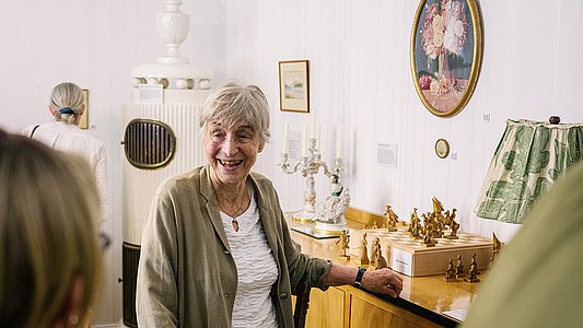 Eine Dame steht lächelnd im rekonstruierten Biedermeierzimmer und legt eine Hand vor einem Schachbrett auf das Buffet