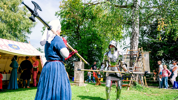 Links im Bild eine Frau im blauen Kleid und weißem Schleier mit einer Hellebarde über der Schulter, die auf einen Kämpfer in Plattenrüstung zuläuft