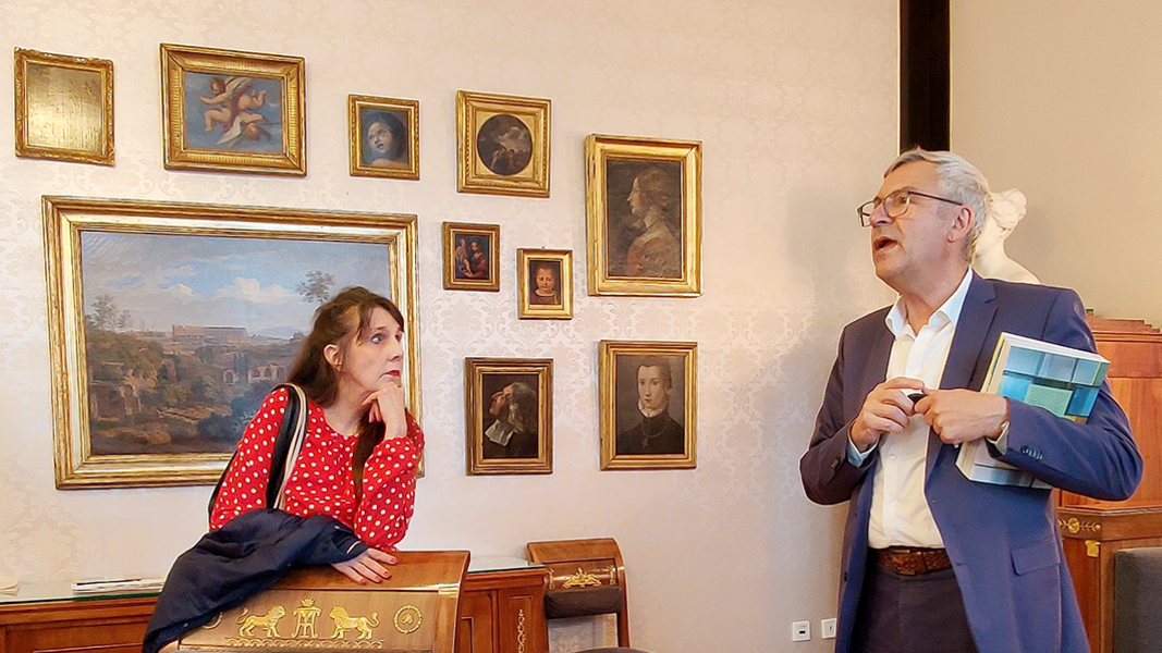 Prof. Dr. Ernst Seidl, mit dem Band "Kunst an der Universität Tübingen", spricht über die an der Wand hängenden, gold gerahmten Gemälde mit Darstellungen Tübingens und universitärer Akteur*innen; links im Bild zuhörend Frau Sabine Schloz