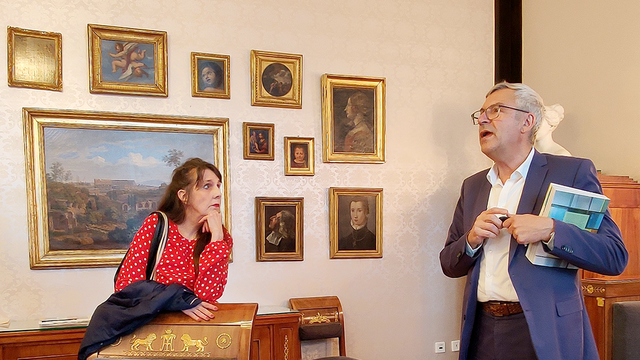 Foto: Serge Le Goff Prof. Dr. Ernst Seidl, mit dem Band "Kunst an der Universität Tübingen", spricht über die an der Wand hängenden, gold gerahmten Gemälde mit Darstellungen Tübingens und universitärer Akteur*innen; links im Bild zuhörend Frau Sabine Schloz
