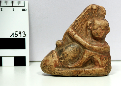 Kleinformatige Steinfigur eines kahlen Mannes (rechts), der einen übergroßen Phallus (links) umarmt; links im Bild ist die Kante eines Maßstabes zu sehen