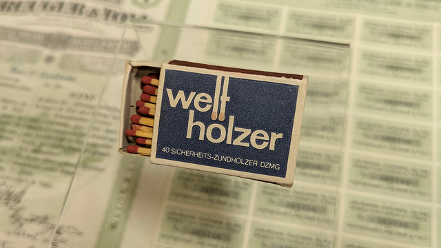 Foto: Universitätsbibliothek Tübingen Eine teils geöffnete Streichholzschachtel mit dem angegilbt-weißen Schriftzug "Welthölzer/40 Sicherheits-Zündhölzer DZMG" vor dunkelblauem Grund; im Hintergrund verwaschen eine aufgeschlagene Zeitung