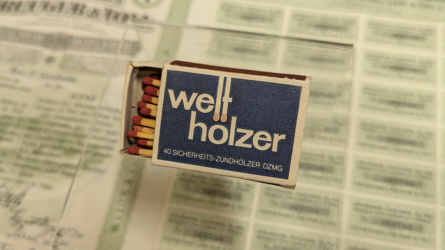 Eine teils geöffnete Streichholzschachtel mit dem angegilbt-weißen Schriftzug "Welthölzer/40 Sicherheits-Zündhölzer DZMG" vor dunkelblauem Grund; im Hintergrund verwaschen eine aufgeschlagene Zeitung