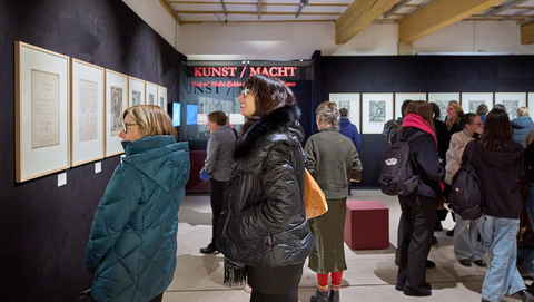 Besuchende drängen sich in der Ausstellung und vor den schwarz bespannten Wänden mit hellen, gerahmten Drucken