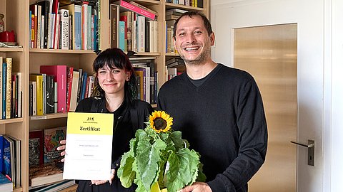 Vor einem Bücherregal und einer weißen Tür stehen lächelnd Tabea Olhorn mit schwarzen Haaren und schwarzer Kleidung, die eine Urkunde mit gelbem Header hochhält, und Michael La Corte in dunklem Pullover, der eine Sonnenblume vor dem Körper hält.