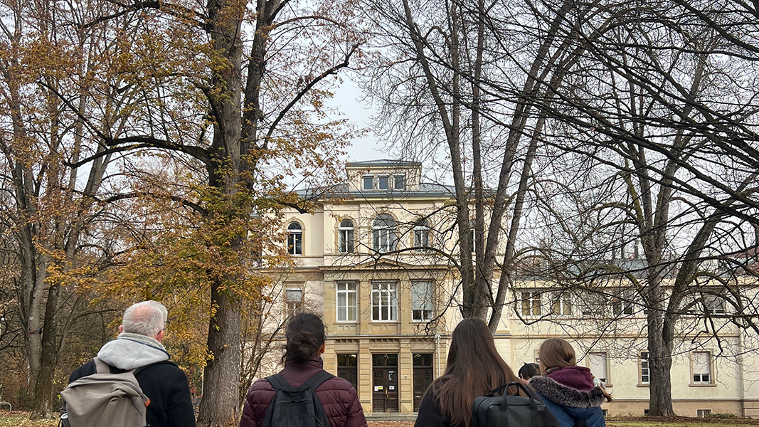 Am unteren Bildrand Kopf- und Schulterbereich der Studierenden in Rückansicht, darüber von kahlen Bäumen zum Teil verdeckt der Haupteingang des Pathologischen Instituts