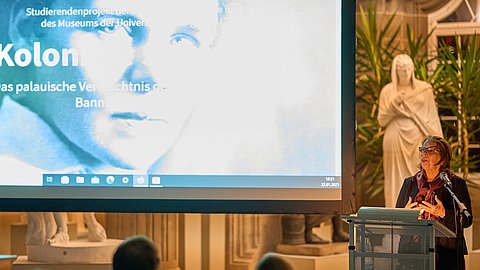 Foto: MUT | Fabian Kurze Prof Dr Gabriele Alex am Rednerpult der Eröffnung „Koloniale Schatten“; auf der Leinwand links das Titelbild der Ausstellung mit dem Schwarz-weiß-Portrait von Elisabeth Krämer-Bannow und dem Titel als Schriftzug