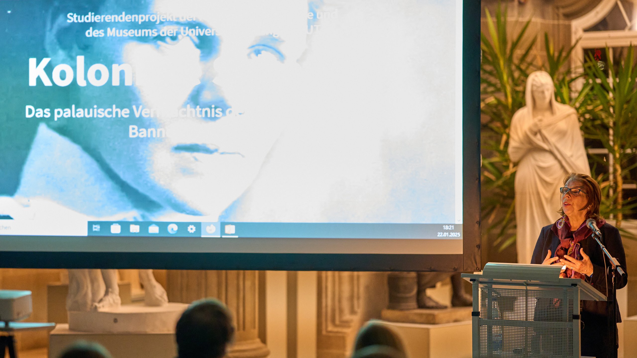 Prof Dr Gabriele Alex am Rednerpult der Eröffnung „Koloniale Schatten“; auf der Leinwand links das Titelbild der Ausstellung mit dem Schwarz-weiß-Portrait von Elisabeth Krämer-Bannow und dem Titel als Schriftzug