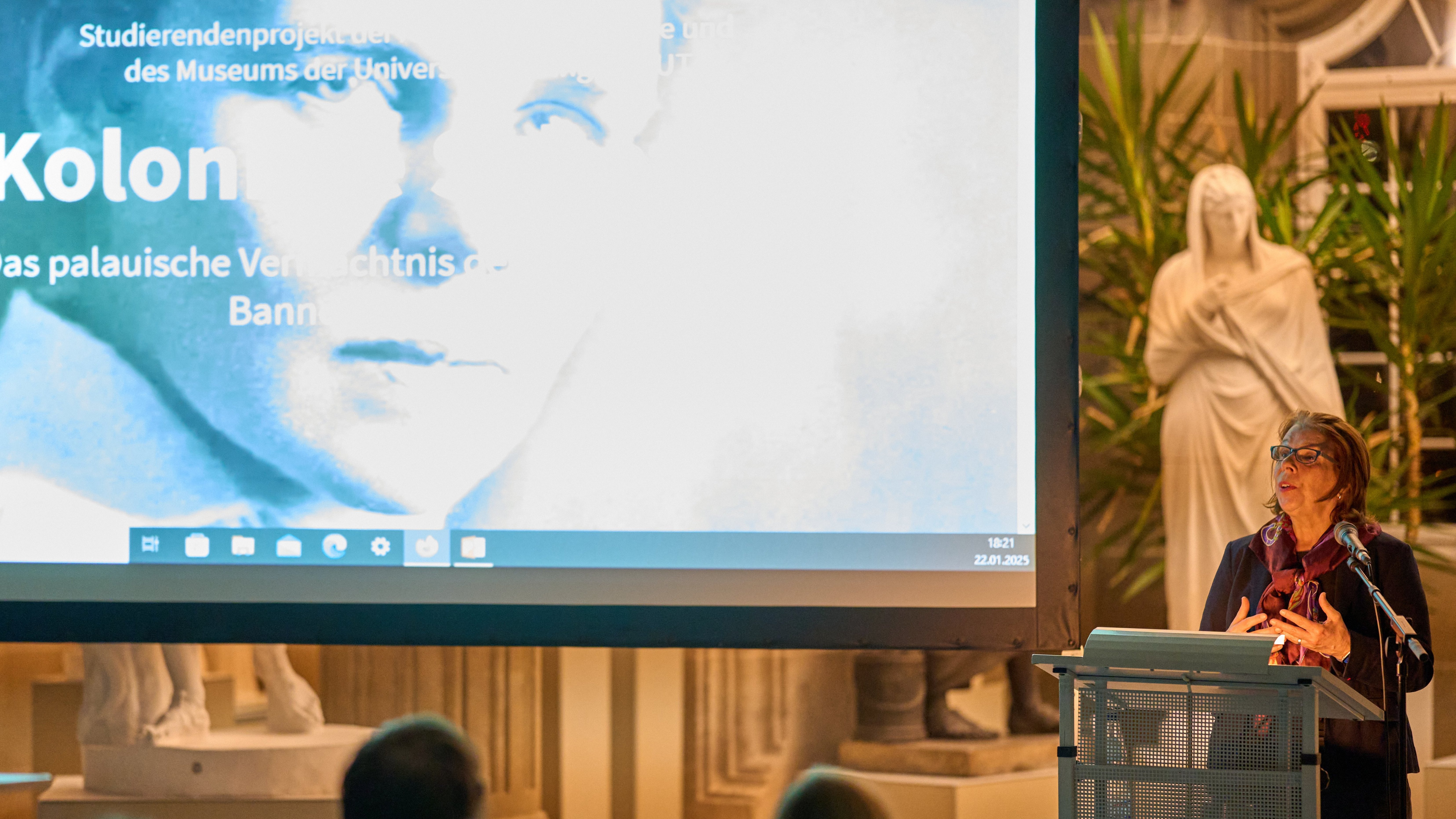 Prof Dr Gabriele Alex am Rednerpult der Eröffnung „Koloniale Schatten“; auf der Leinwand links das Titelbild der Ausstellung mit dem Schwarz-weiß-Portrait von Elisabeth Krämer-Bannow und dem Titel als Schriftzug