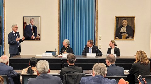 Foto: MUT | Ernst Seidl Am Rednertisch des Großen Senats der Uni Tübingen sitzen vor blauem Vorhang mehrere Personen und blicken zum links hängenden Portrait von Bernd Engler