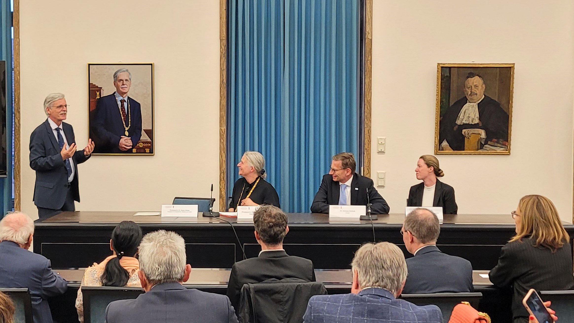 Am Rednertisch des Großen Senats der Uni Tübingen sitzen vor blauem Vorhang mehrere Personen und blicken zum links hängenden Portrait von Bernd Engler