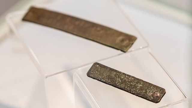 Auf zwei schräg stehenden Acrylbrücken liegen schräg hintereinander zwei längliche Bronzetäfelchen mit gepunzter beziehungsweise geritzter Inschrift