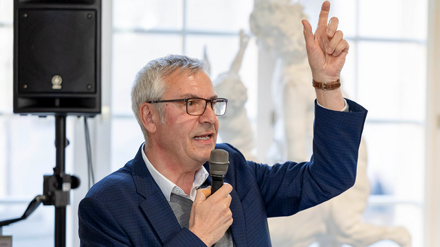 Prof. Dr. Ernst Seidl, ein grauhaariger Mann im blauen Anzug mit gestikulierend erhobener Linker, spricht nach rechts gewandt in ein Mikrophon in seiner rechten Hand, im Hintergrund ein sonnendurchleuchtetes Fenster sowie links eine Lautsprecherbox