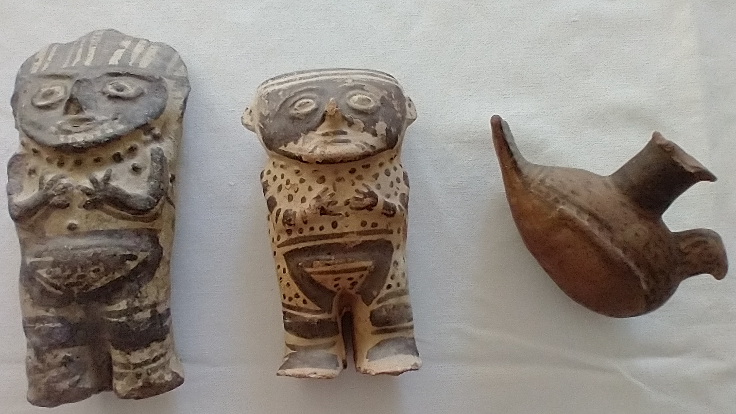 Drei altamerikanische Keramikobjekte auf grauem Seidenpapier: Links zwei humanoide Figuren mit flachen Gesichtern und schwarzfiguriger Bemalung, rechts ein Gefäß in Form eines sitzenden Vogels mit der Tülle auf dem Rücken