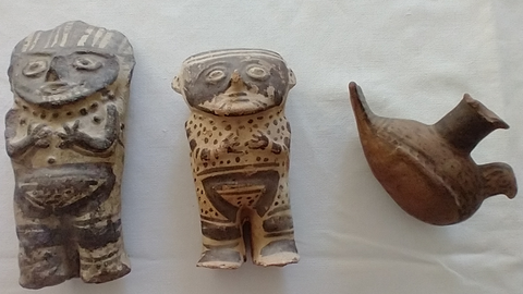 Drei altamerikanische Keramikobjekte auf grauem Seidenpapier: Links zwei humanoide Figuren mit flachen Gesichtern und schwarzfiguriger Bemalung, rechts ein Gefäß in Form eines sitzenden Vogels mit der Tülle auf dem Rücken