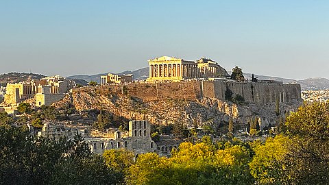 Fernansicht der Athener Akropolis, vom links von der gelblichen Abendsonne angestrahlt, mit Baumkronen im Vordergrund
