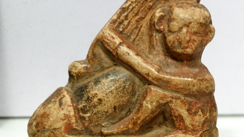 Kleinformatige Steinfigur eines kahlen Mannes (rechts), der einen übergroßen Phallus (links) umarmt