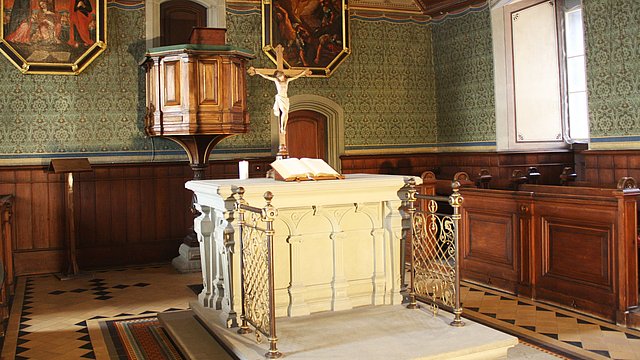 Foto: MUT Blick in den Altarbereich der Schlosskirche mit weißem Altar mit aufgeschlagener Bibel, gekreuzigter Christusfigur und hinaufführender Treppe, im Hintergrund Kanzel, flankiert von Malereien des gekreuzigten (links) und in den Himmel fahrenen Christus (rechts)