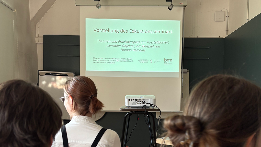 Titelfolie des Vortrags "Vorstellung des Exkursionsseminars. Theorien und Praxisbeispiele zur Ausstellbarkeit "sensibler Objekte", am Beispiel von Human Remains"; im Vordergrund Zuschauende, die sich erwartungsvoll nach links und rechts wenden