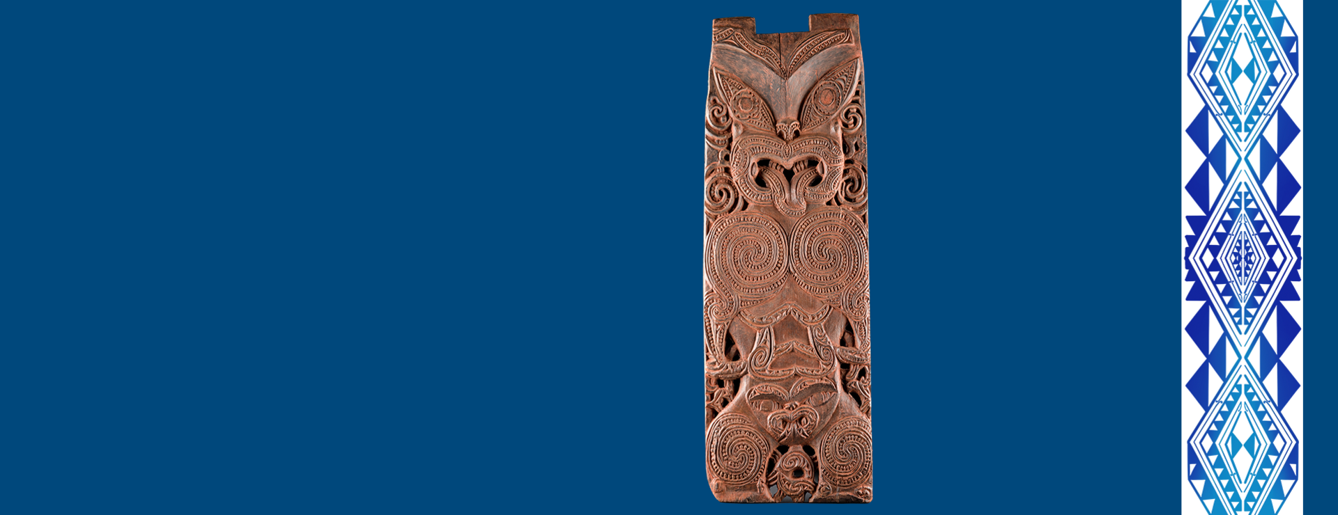 Geschnitztes Holzpaneel (pou) mit der Darstellung einer Ahnin der Te Aitanga-a-Hauiti vor dunkelblauem Hintergrund; rechts ein schmaler Streifen mit einem vor allem aus dreieckigen Formen zusammengesetzten, abstrakten, blauen Design vor weißem Hintergrund