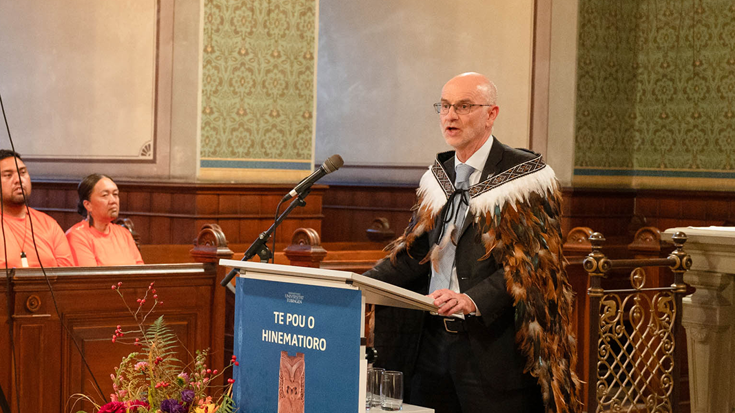 Am Rednerpult mit dem blauen Ausstellungsplakat von "Te Pou o Hinematioro" steht der neuseeländische Botschafter Craig John Hawke im Anzug und zeremoniellem Federumhang