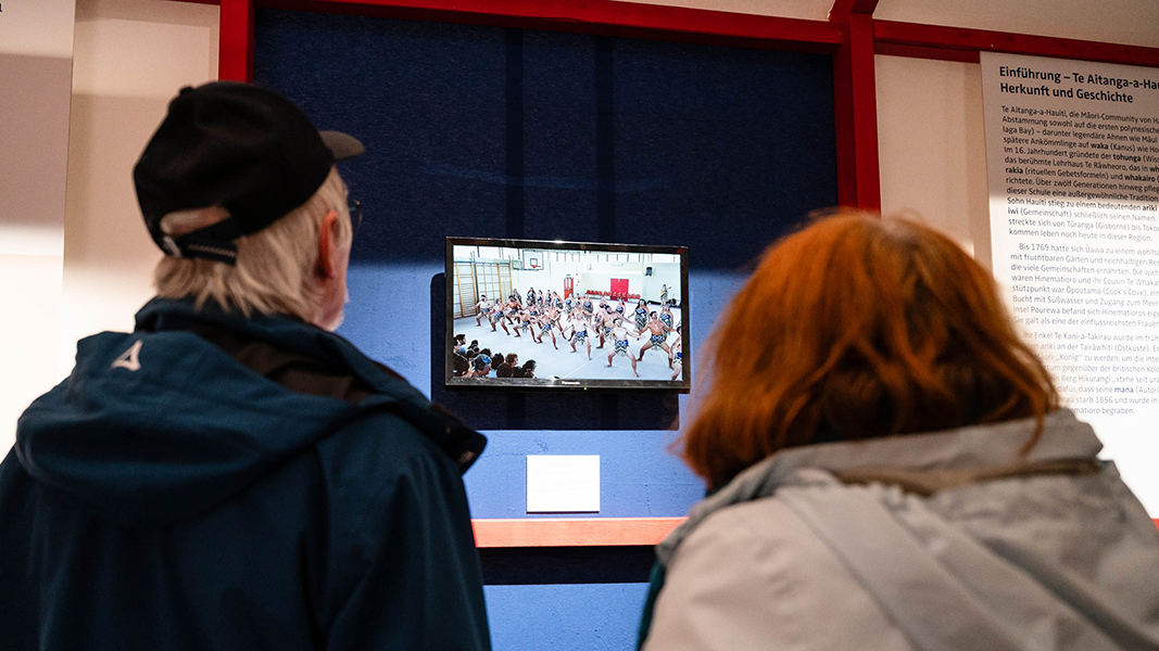 Zwei Besuchende, von hinten gesehen, betrachten eine Videovorführung in der Ausstellung; der Bildschirm zeigt eine Gruppe Maori während eines Tanzes in tiefer, breitbeiniger Haltung