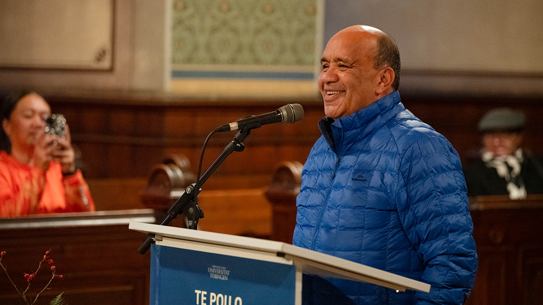 Am Rednerpult mit dem blauen Ausstellungsplakat von "Te Pou o Hinematioro" steht lächelnd Victor Walker, Sprecher der Delegation der Te Aitanga-a-Hauiti; links im Hintergrund ist ein Mitglied der Delegation mit zum Fotografieren erhobenen Smartphone zu sehen