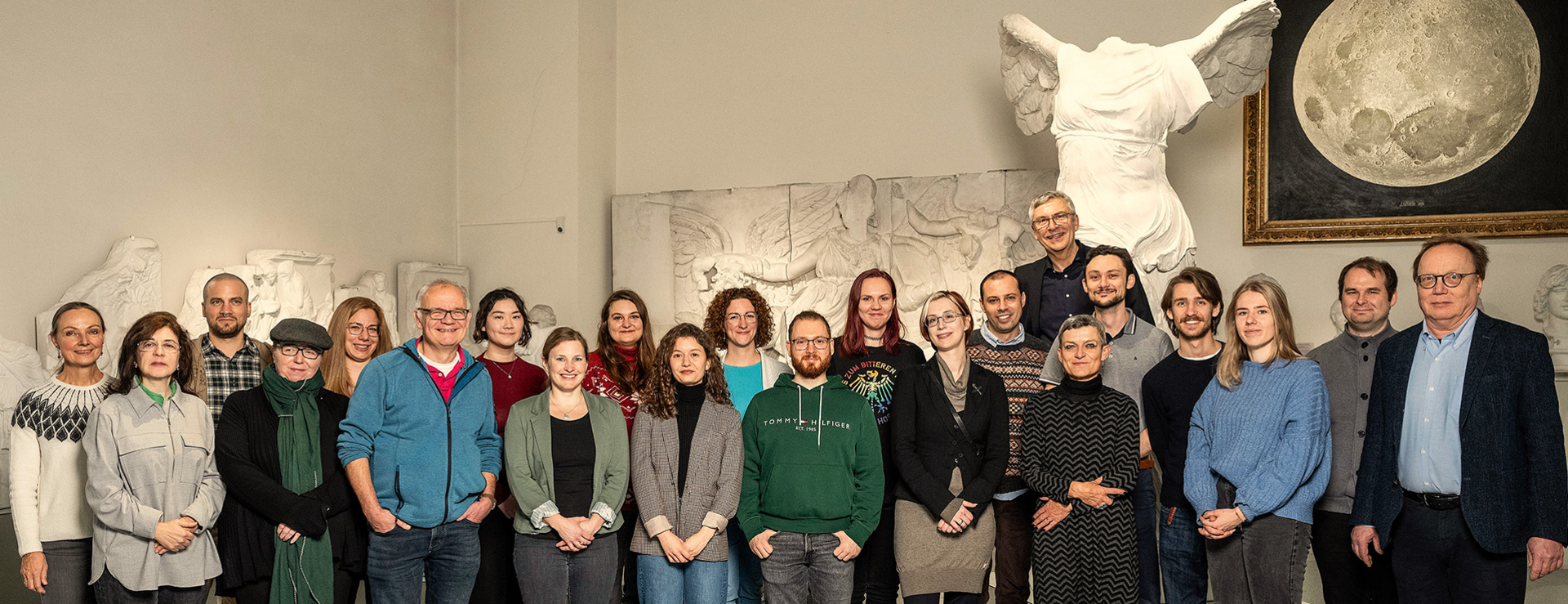 Foto: MUT | V. Marquardt Teamfoto der MUT-Mitarbeitenden im Halbkreis im Rittersaal auf Schloss Hohentübingen; im Hintergrund der Gipsabguss der Nike von Samothrake und das realistische Mond-Gemälde