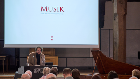 Dr. Jörg Büchler am Rednerpult, hinter ihm die Titelfolie "Musik in den Sammlungen der Universität Tübingen" auf der Leinwand des Pfleghofsaals