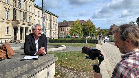 Prof. Dr. Ernst Seidl (mittig) spricht vor dem Hintergrund der Neuen Aula in der Münzgasse in die Kamera, die von Alexander Ries (rechts, Rückenansicht) gehalten wird.