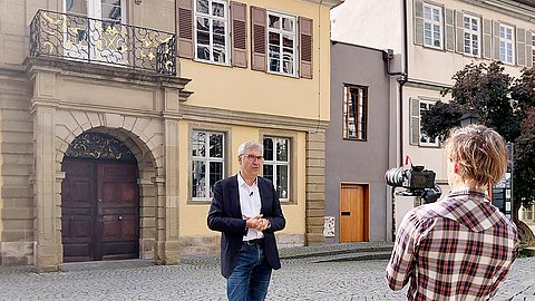 Prof. Dr. Ernst Seidl (mittig) spricht vor dem Hintergrund der Alten Aula in der Münzgasse in die Kamera, die von Alexander Ries (rechts, Rückenansicht) gehalten wird.