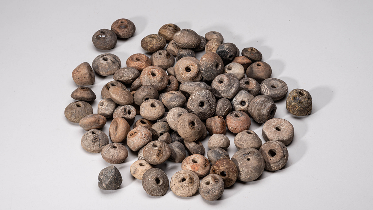 Spinnwirtel und Webgewichte mit eingeritzten Linien, in der Bildmitte angehäuft, vor neutral-grauem Hintergrund.