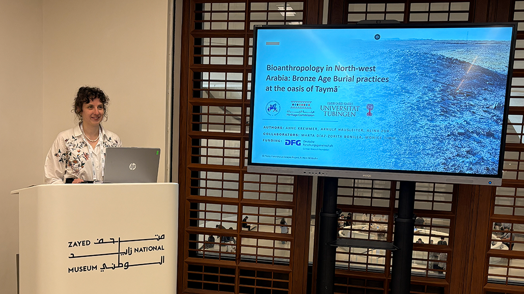 Dr. Anne Kremmer am weißen Rednerpult mit ihrer Präsentation mit dem Titel "Bioanthropology in North-west Arabia: Bronze Age Burial practices at the oasis of Taymā"" auf einem Bildschirm rechts